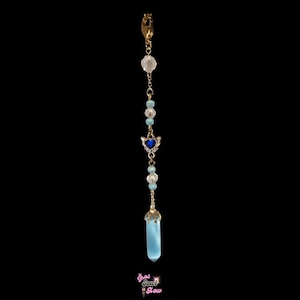 May include: A gold-colored charm with a clip, featuring a series of beads in white and light blue, a silver-colored heart with blue accents, and a light blue crystal pendant. The charm is suspended against a black background.