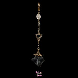 May include: A gold-colored belly button ring with a clear crystal bead, a heart-shaped charm with wings and a dark blue gem, and a faceted dark crystal pendant. The ring is suspended on a delicate chain against a black background.