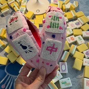 Puede incluir: Zapatillas de tela rosa con diseños de cuentas. Las zapatillas tienen un interior rojo y están decoradas con cuentas blancas y patrones bordados coloridos. El fondo incluye fichas de Mahjong amarillas y blancas.