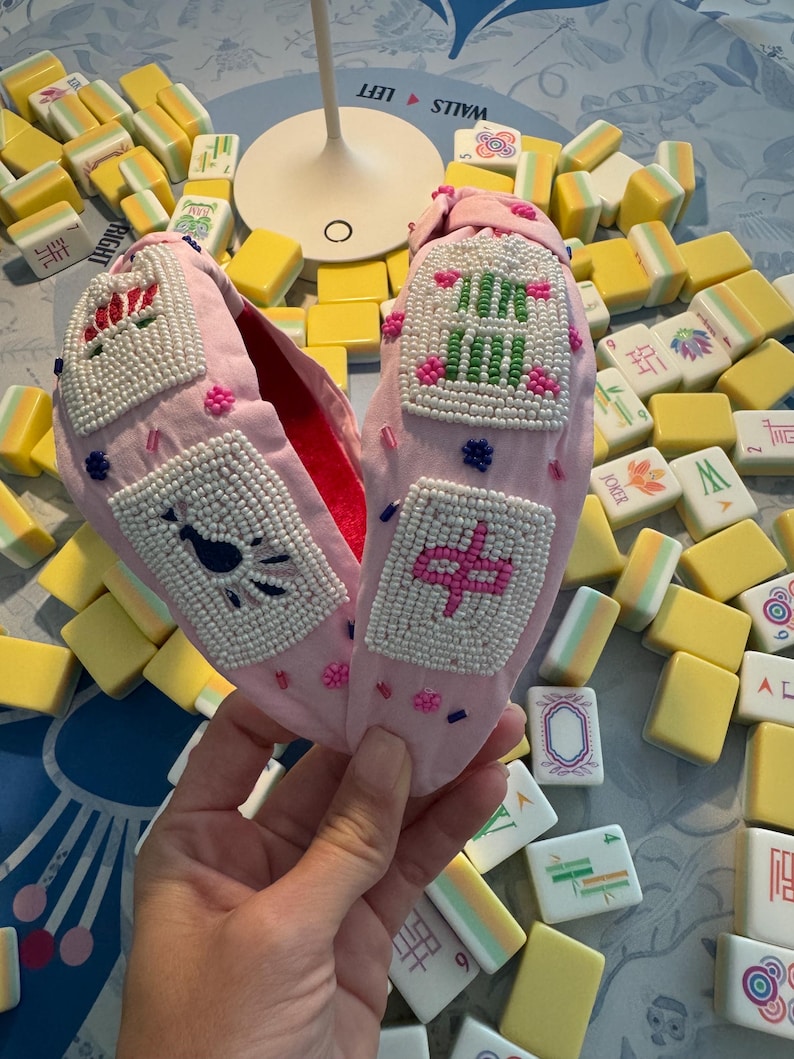 Puede incluir: Zapatillas de tela rosa con dise&ntilde;os de cuentas. Las zapatillas tienen un interior rojo y est&aacute;n decoradas con cuentas blancas y patrones bordados coloridos. El fondo incluye fichas de Mahjong amarillas y blancas.