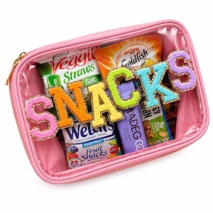 May include: A rectangular pink clear-front pouch filled with various snack packages including Veggie Straws, Welch's fruit snacks, Goldfish, and a MadeGood granola bar. The word SNACKS is spelled out in large, colorful, beaded varsity-style letters.