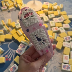 Puede incluir: Una diadema rosa claro con adornos de cuentas. La diadema presenta dos dise&ntilde;os rectangulares con cuentas, uno con un p&aacute;jaro y otro con una corona. Peque&ntilde;as cuentas rosas y detalles florales dispersos. El fondo incluye fichas de Mahjong amarillas y blancas.