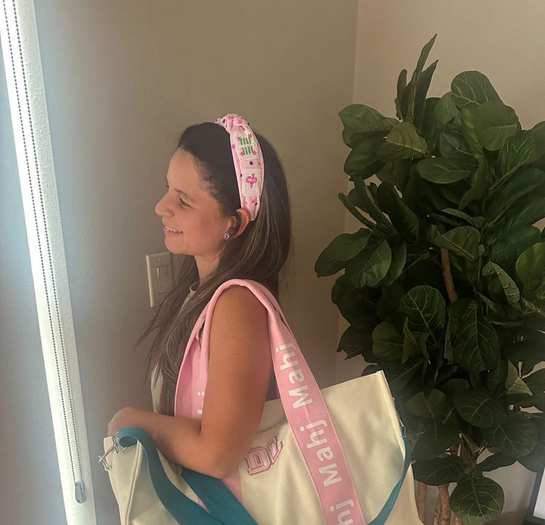Puede incluir: Una diadema rosa claro con un patr&oacute;n repetido de texto y peque&ntilde;os gr&aacute;ficos. Un bolso tote blanco con una correa rosa que tiene el texto "Mahj Mahj" impreso. La bolsa tiene una correa azul turquesa.