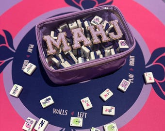 May include: A purple clear plastic case with a zipper closure filled with mahjong tiles. The case has the word "MAHJ" spelled out in pink glitter letters on the front. The case is sitting on a blue and pink mahjong game mat.