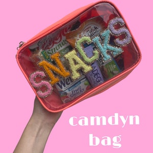 May include: A clear plastic bag with a coral pink zipper and a white zipper pull. The bag is filled with various snacks, including fruit snacks, granola bars, and crackers. The bag has the word "SNACKS" spelled out in colorful, glittery letters.