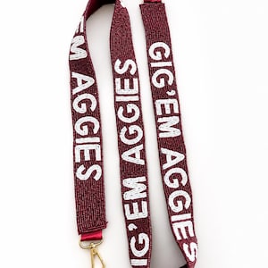 Könnte beinhalten: Ein kastanienbrauner, perlenbesetzter Taschengurt mit dem weißen Schriftzug GIG 'EM AGGIES, der sich über die gesamte Länge wiederholt. Der Gurt ist an beiden Enden mit goldfarbenen Karabinerverschlüssen aus Metall versehen.