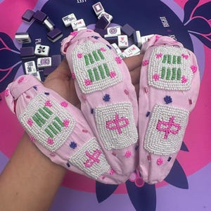 Puede incluir: Soportes de tela rosa para fichas de mahjong con dise&ntilde;os de cuentas en blanco, verde y rosa. Los soportes presentan caracteres chinos y patrones geom&eacute;tricos. El fondo incluye fichas de mahjong y un tapete de juego morado y rosa.