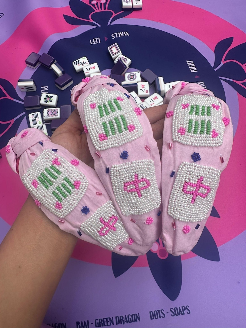 Puede incluir: Soportes de tela rosa para fichas de mahjong con dise&ntilde;os de cuentas en blanco, verde y rosa. Los soportes presentan caracteres chinos y patrones geom&eacute;tricos. El fondo incluye fichas de mahjong y un tapete de juego morado y rosa.
