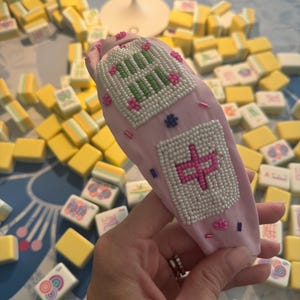 Puede incluir: Una diadema rosa claro con dise&ntilde;os de cuentas, incluyendo caracteres chinos y patrones florales. La diadema est&aacute; rodeada de fichas de Mahjong amarillas y blancas, sugiriendo un tema de juego.