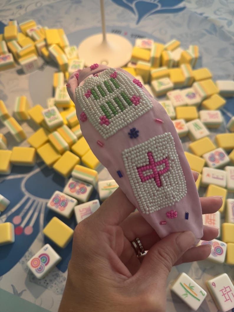 Puede incluir: Una diadema rosa claro con dise&ntilde;os de cuentas, incluyendo caracteres chinos y patrones florales. La diadema est&aacute; rodeada de fichas de Mahjong amarillas y blancas, sugiriendo un tema de juego.