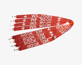 Puede incluir: Una colección de cordones de cuentas naranjas con el texto blanco HOOK 'EM y símbolos de cruz blancos. Cada correa está terminada con un mosquetón giratorio de metal plateado en el extremo, diseñado para sujetar objetos a la correa de tela.