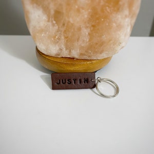 May include: A brown leather keychain with the name "JUSTIN" embossed on it. The keychain is attached to a silver metal ring. The keychain is on a white surface, with a large, light orange Himalayan salt lamp in the background.