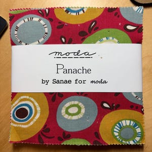 May include: A stack of fabric squares with a floral pattern in red, yellow, blue, and green. The fabric is wrapped with a white band that reads "moda Panache by Sanae for moda."