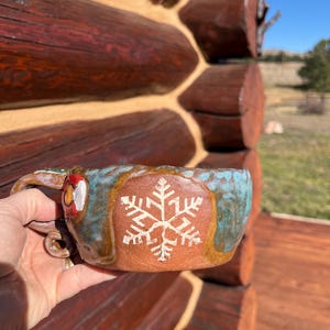 May include: A handmade ceramic mug with a turquoise and brown glaze. The mug features a white snowflake design and a unique handle. The mug is held in front of a rustic log cabin.