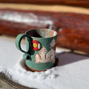 May include: A teal ceramic mug with a handle, featuring a Colorado state flag emblem and a relief design of trees and a cabin. The mug is set in the snow, with a wooden background.