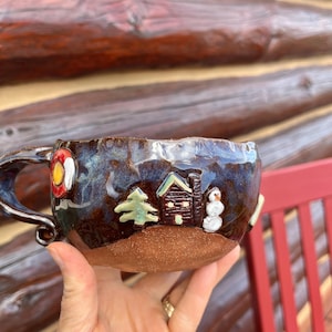 May include: A handmade ceramic mug with a dark blue glaze and a brown base. The mug features a winter scene with a cabin, snowman, and tree. A red and yellow life preserver is on the side.