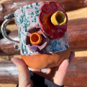 May include: A handmade ceramic mug with a unique floral design. The mug features a speckled blue and white glaze with green accents. Two vibrant, three-dimensional flowers in red, purple, and yellow adorn the mug's exterior. The base is a warm terracotta color.