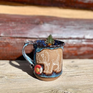 May include: A handmade ceramic mug with a brown bear design. The mug has a blue and brown glaze, a small tree inside, and a red and yellow life preserver detail. The mug is sitting on a wooden surface.