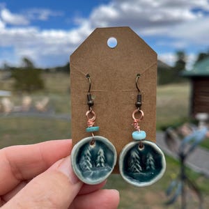 May include: Handmade ceramic earrings featuring a forest scene in a teal glaze. The earrings have copper wire accents and turquoise beads. The earrings are attached to a brown tag.
