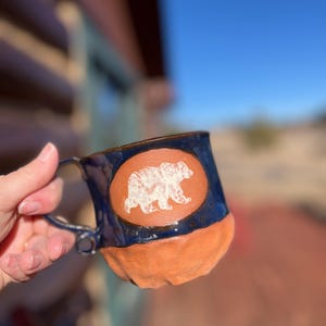 May include: Handmade ceramic mug with a blue glazed top and orange base. A white bear silhouette is centered on an oval orange patch. The mug has a curved handle and is held by a person.