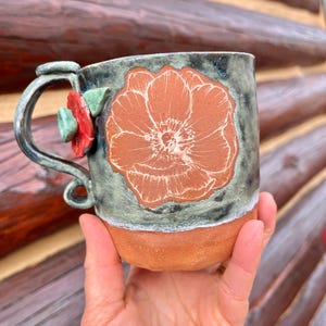 May include: Handmade ceramic mug with a large orange flower design. The mug has a gray-green glaze with a terracotta-colored base. A red and green flower adorns the handle, adding a touch of whimsy to this unique piece.