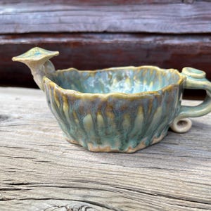 May include: A handmade ceramic bowl with a unique mushroom-shaped spout and handle. The bowl features a textured, organic design with a blend of teal, brown, and yellow-green glazes. The bowl is approximately 6 inches wide and 3 inches tall.