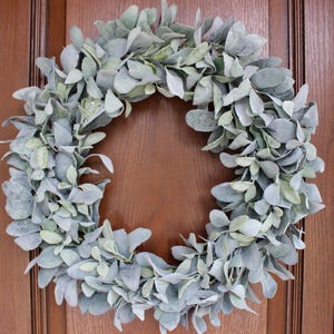 May include: A wreath made of artificial lambs ear leaves. The leaves are a light green colour and have a fuzzy texture. The wreath is hanging on a wooden door.