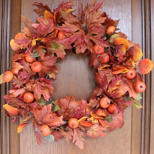 Peut inclure: Une couronne d'automne circulaire composée d'un arrangement dense de feuilles d'érable artificielles dans des tons de rouge profond, orange brûlé et jaune doré, agrémentée de petites citrouilles orange et de grappes de baies jaunes.