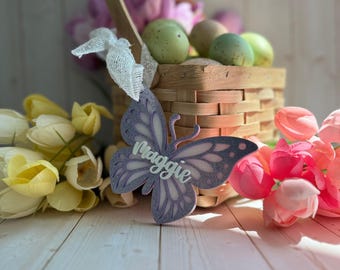May include: Purple butterfly-shaped Easter decoration with the name "Maggie" written in white lettering. The decoration is sitting on a white wooden surface with a basket of Easter eggs and flowers in the background.