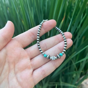 May include: Silver beaded bracelet with turquoise beads and a silver letter "B" charm. The bracelet is held in a hand against a backdrop of green leaves. A delicate piece of jewellery.