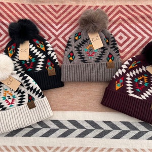 May include: Four knitted winter hats with pom-poms and geometric patterns. The hats are in black, grey, white, and burgundy with colourful accents. Each hat has a brown leather patch with the letters "C.C." on it.