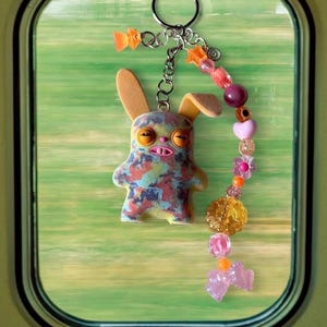 Peut inclure: Un porte-clés représentant une petite figurine de lapin en peluche avec un motif camouflage multicolore, des yeux orange et deux dents de devant proéminentes. Un cordon de perles roses, orange et jaunes est attaché à l'anneau métallique.