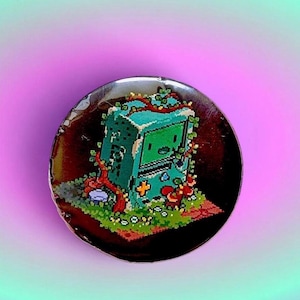 May include: A circular pin featuring a pixel art illustration of a teal, rectangular, box-like character with a screen face, surrounded by green grass, small white flowers, and red vines, set against a dark brown background within the pin.