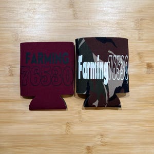 May include: Two beverage can coolers. One is maroon with the word "FARMING" and the number "76530" in black. The other is camouflage with the same text in white.