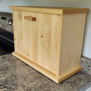Könnte beinhalten: Eine Brotbox aus Holz mit schlichtem Design. Die Box ist aus hellem Holz gefertigt und hat einen kleinen Holzgriff. Die Box steht auf einer Arbeitsplatte.