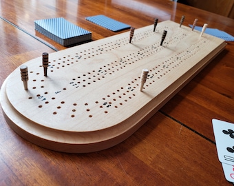May include: A wooden cribbage board with pegs in the holes. The board is made of light wood and has a rounded edge. There are playing cards on the table next to the board.