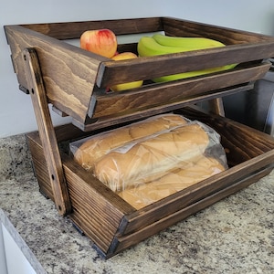 Puede incluir: Una cesta de fruta y pan de madera de dos niveles con un tinte marrón oscuro. El nivel superior contiene plátanos y manzanas, mientras que el nivel inferior contiene barras de pan en bolsas de plástico.