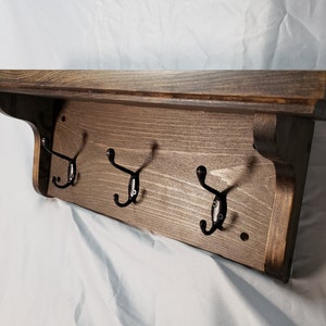 May include: A wooden shelf with three black metal hooks. The shelf is stained a dark brown color and has a slightly distressed look. The hooks are attached to the front of the shelf and are spaced evenly apart.
