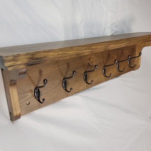 May include: A wooden shelf with six black metal hooks. The shelf is stained a dark brown color and has a slightly distressed finish. The shelf is mounted to a wall.