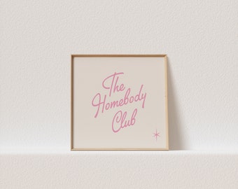 May include: A framed print with a light beige background and a pink inscription that reads "The Homebody Club" with a small pink star in the bottom right corner.
