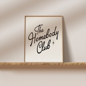 May include: A framed print with the text "The Homebody Club" in a black cursive font with a small star symbol.