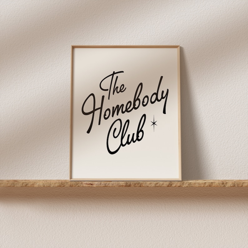 May include: A framed print with the text "The Homebody Club" in a black cursive font with a small star symbol.