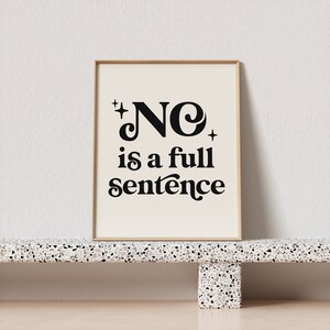 May include: A framed print with the words "NO is a full sentence" in black lettering. The print has a cream-colored background and is in a light brown frame. The print is displayed on a speckled shelf.