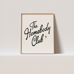 May include: A framed print with a white background and black text that reads "The Homebody Club" with a small star next to the word "Club".