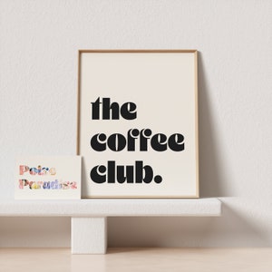 May include: A framed print with the text "the coffee club." in black bold letters on a cream background. A small card with the text "Poire Paradise" in colorful letters is in the foreground.