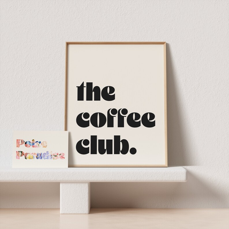 May include: A framed print with the text "the coffee club." in black bold letters on a cream background. A small card with the text "Poire Paradise" in colorful letters is in the foreground.