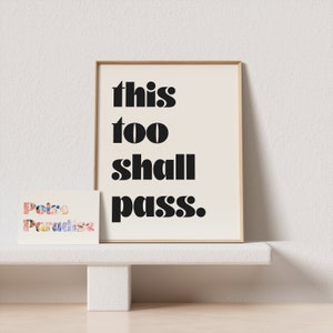 K&ouml;nnte beinhalten: Ein gerahmter Druck mit dem Text "this too shall pass." in schwarzen Fettdruck auf cremefarbenem Hintergrund. Der Druck steht auf einem wei&szlig;en Regal.