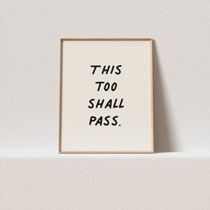 Puede incluir: Una impresión enmarcada con el texto "THIS TOO SHALL PASS." en fuente manuscrita negra sobre fondo blanco.