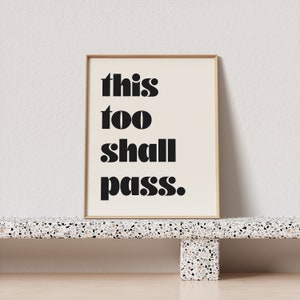 K&ouml;nnte beinhalten: Ein gerahmter Druck mit dem Text "this too shall pass." in schwarzen Fettdruck auf cremefarbenem Hintergrund.
