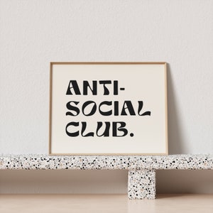 May include: A framed print with the text "ANTI-SOCIAL CLUB." in black stylized lettering on a white background. The frame is a light wood color and the print is sitting on a terrazzo shelf.